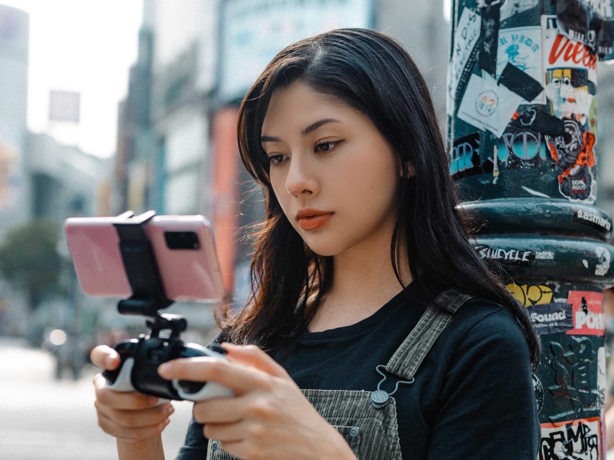 A young woman playing an Xbox game on her mobile phone, while holding a controller and leaning against a lamp post.