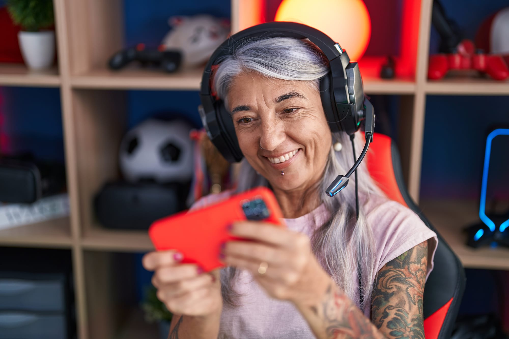 Smiling older woman with silver hair and tattoos wearing gaming headset, holding red controller in home gaming setup, representing mature female gamers over 50